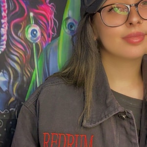 Puede incluir: Chaqueta vaquera y gorra de camionero negras con la palabra "REDRUM" bordada en rojo. La gorra es de estilo camionero de malla. La chaqueta tiene cierre de botones y cuello. El fondo presenta un mural colorido.