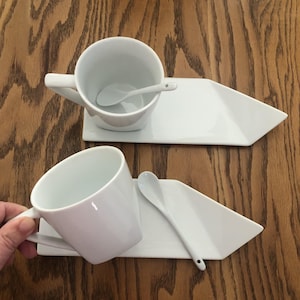 May include: Two white ceramic coffee cups with matching saucers and spoons. The cups have a handle and sit on a geometric-shaped saucer. A small spoon rests inside each cup. The set is on a wooden surface.