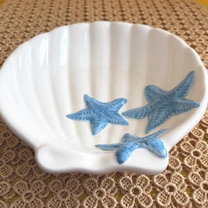 May include: A white ceramic bowl shaped like a scallop shell, featuring three raised, textured blue starfish attached to the interior surface. The bowl has a glossy finish and a fluted design that mimics the natural ridges of a seashell.