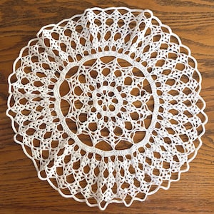 May include: A circular white crochet doily featuring an intricate openwork pattern. The design consists of a central star-like motif surrounded by concentric rings of delicate, interconnected loops and stitches, finished with a scalloped edge.