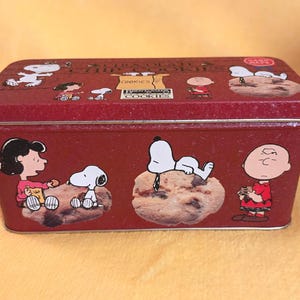 May include: A rectangular red metal tin featuring Peanuts characters including Snoopy, Charlie Brown, and Lucy. The lid and sides display illustrations of the characters with chocolate chip cookies and the text Chocolate Chip Cookies and Brent & Sam's Cookies.