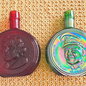 May include: Two vintage glass bottles with stoppers. One is red with Thomas Jefferson's portrait, the other is iridescent with Dwight D. Eisenhower's portrait. Both bottles have a circular shape and are likely decorative.
