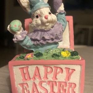 May include: A decorative Easter figurine featuring a white bunny wearing a blue hat and outfit, holding an egg. The figurine is in a pink box with "HAPPY EASTER" in white letters. The bunny is surrounded by green grass and flowers.