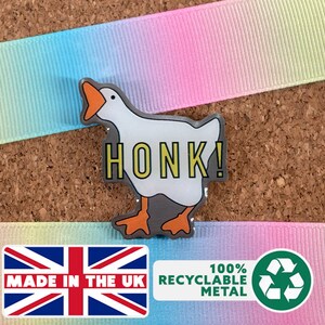 Puede incluir: Pin metálico con un ganso blanco con pico y patas naranjas, y la palabra "HONK!" en amarillo. El pin está sobre un fondo de corcho con una cinta arcoíris. Fabricado en metal 100% reciclable.