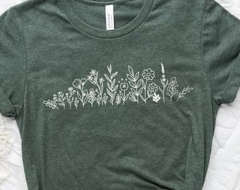 May include: Dark green t-shirt with a white floral design across the chest. The shirt is knotted at the bottom. A neutral-toned hat, a cup of coffee, and a book are also visible in the image.