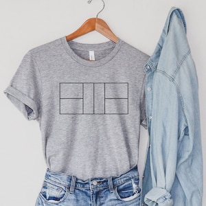 Peut inclure: Un t-shirt gris avec un motif noir de court de tennis sur le devant. Le t-shirt est suspendu à un cintre en bois et est associé à une chemise en jean bleu clair.