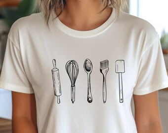 May include: White t-shirt with a graphic of five kitchen utensils: a rolling pin, whisk, spoon, pastry brush, and spatula.