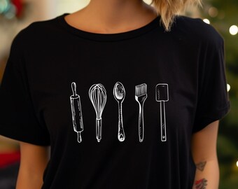 May include: A woman wearing a black t-shirt with a white graphic of kitchen utensils, including a rolling pin, whisk, spoon, brush, and spatula.