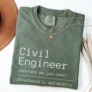 Puede incluir: Camiseta verde oliva con las palabras "Civil Engineer" en blanco, una guía de pronunciación y la frase "Structurally optimistic." Un collar dorado con un colgante descansa sobre la camiseta doblada sobre una superficie blanca.