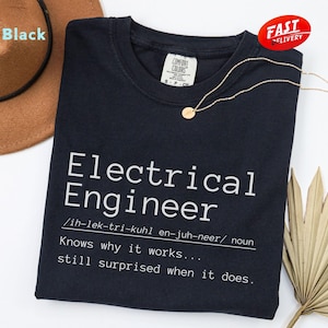 May include: A black t-shirt with the white text "Electrical Engineer", a definition, and a humorous quote. A gold necklace and a brown hat are also visible. The shirt has a "Fast Delivery" sticker.