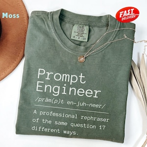 May include: A moss green t-shirt with the text "Prompt Engineer" in white. The shirt includes a definition and is paired with a gold necklace. A "Fast Delivery" sticker is visible.