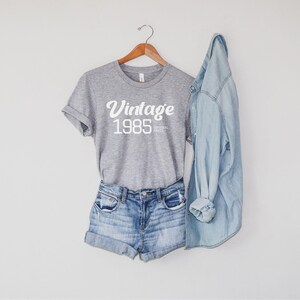 May include: A gray t-shirt with the text "Vintage 1985" in white. The shirt is hanging on a wooden hanger and is paired with a light blue denim jacket and denim shorts.