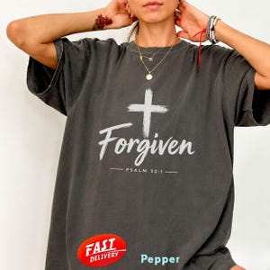 May include: A charcoal grey short-sleeve t-shirt featuring a white graphic of a cross above the word Forgiven and the text PSALM 32:1. The shirt has a relaxed fit with a crew neckline and a faded, vintage-inspired wash throughout the fabric.