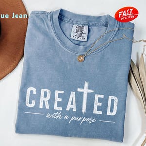 Op de afbeelding: Een blauw spijkerbroekkleurig t-shirt met de witte woorden "CREATED with a purpose". Het shirt heeft een ronde hals en een klein label op de kraag. Een gouden ketting met een hanger ligt op het shirt.