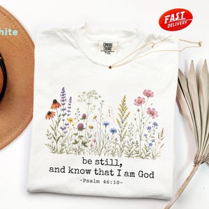 May include: White t-shirt with a floral design featuring wildflowers in various colours. The shirt has the text "be still, and know that I am God -Psalm 46:10-". A gold necklace with a star pendant is on the shirt.