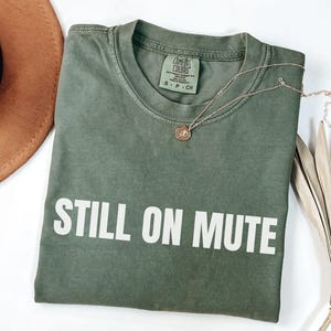May include: An olive green t-shirt with the words "STILL ON MUTE" printed in white. A gold necklace with a small pendant rests on the shirt. A brown hat and dried plant are in the background.