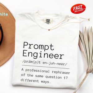 以下が含まれることがあります： 「Prompt Engineer」の黒い文字がプリントされた白いTシャツ。発音記号と定義も含まれています。星のペンダントが付いたゴールドのネックレスがシャツの上にかけられています。「Fast Delivery」のステッカーが右上隅にあります。