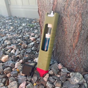 May include: An olive green and red device with a rectangular body and a spring mechanism. The device has a clear tube containing a blue liquid. A black square cap is next to the device. The device is resting on a bed of rocks.