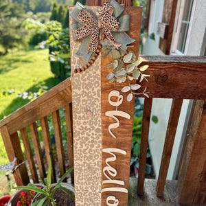 May include: Wooden welcome sign with a leopard print design and the words "oh hello" in white script. The sign is adorned with a bow featuring leopard print and sage green fabric, along with faux greenery and wooden beads. The sign is displayed on a wooden porch.