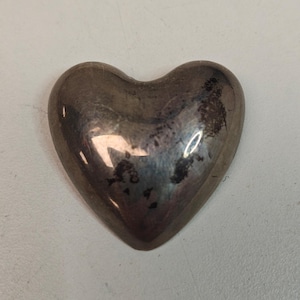 May include: A silver-coloured heart-shaped object with a slightly tarnished surface. The heart has a smooth, rounded shape and reflects light, showing some dark spots. The object is likely made of metal.