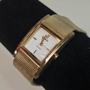 May include: A gold-toned Timex watch with a rectangular face and a mesh band. The watch face is white with black hands and the word "INDIGLO" is printed at the bottom. The watch is displayed on a black surface.