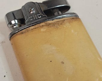 May include: A vintage lighter with a cream-colored body and silver-toned metal accents. The lighter has a rectangular shape with rounded edges and a hinged top. The text "CMC" is visible on the top. The lighter shows signs of wear.