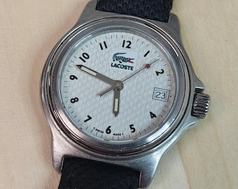 May include: A silver-toned Lacoste watch with a white face featuring the brand's logo and a date display. The watch has a black fabric band. The watch face displays the numbers 1 through 12, with a second hand and minute and hour hands.