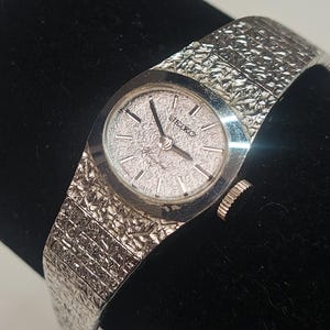 May include: A vintage Seiko watch with a silver textured strap and a round face. The watch face is silver with black hour markers and hands. The watch is displayed on a black surface.