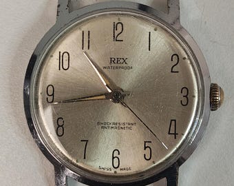 May include: A vintage REX wristwatch with a silver-colored face and a stainless steel case. The watch face displays black numerals, hands, and the text "REX WATERPROOF SHOCKRESISTANT ANTIMAGNETIC SWISS MADE". The watch has a gold-colored crown.