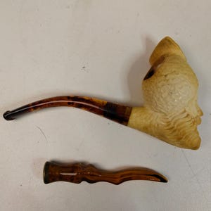 May include: A tobacco pipe with a cream-colored carved bowl featuring a detailed face and a curved, translucent brown tortoiseshell-patterned stem. Below the pipe lies a separate, matching brown tortoiseshell-patterned stem piece with a wavy shape.