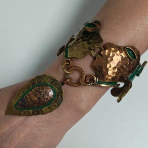 Op de afbeelding: Een gehamerde messing armband met een blad-vormige bedel en kikkerachtige figuren. De armband heeft groene emaille accenten en een koperkleurige afwerking. De armband is een uniek sieraad.