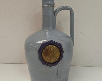 May include: A glossy light gray ceramic jug with a rounded body, a narrow neck, and a single handle. A circular gold-colored medallion with an embossed profile portrait is centered on the front, framed by a dark purple wax-style seal design.