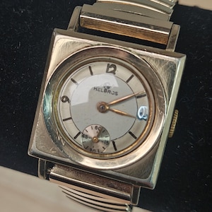 Op de afbeelding: Een goudkleurig Helbros polshorloge met een vierkante wijzerplaat en een ronde wijzerplaat. Het horloge heeft een kleine seconden subdial en een datumvenster. De horlogeband is van flexibel, goudkleurig metaal. De wijzerplaat heeft de nummers 12, 9 en 3.
