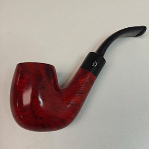 May include: A red and black tobacco pipe with a curved stem. The bowl is a deep red with a swirling pattern, and the stem is black. A black band with a white crown logo is on the pipe.