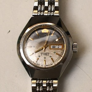 May include: A vintage silver Orient automatic wristwatch with a silver dial and a date display. The watch has a stainless steel strap and a black bezel. The watch face displays the day and date.