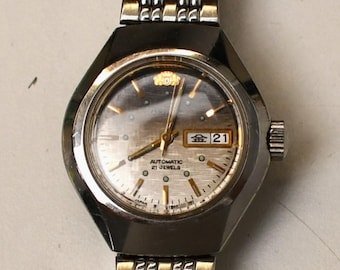 May include: A vintage silver-toned Orient automatic wristwatch with a silver dial and a date display. The watch has a stainless steel band and a black bezel. The watch face displays the day and date.