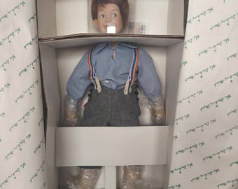 May include: A porcelain doll of a young boy, packaged in a box. The doll has red hair and is wearing a blue striped shirt with rainbow suspenders and gray pants. The doll's arms and legs are wrapped in bubble wrap. The box is labeled "The Danbury Mint."