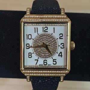 May include: A gold-toned rectangular wristwatch with a white face and black band. The watch face features a sparkling, circular design in the center and is surrounded by a bezel set with small, clear stones. The watch band is black and textured.