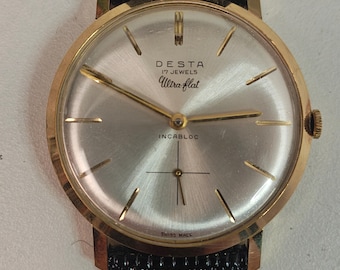 May include: A gold-toned wristwatch with a silver face and black leather band. The watch face features the brand name "DESTA", the words "Ultra flat", and "17 Jewels". The watch also has the word "INCABLOC" and "SWISS MADE" on the face.