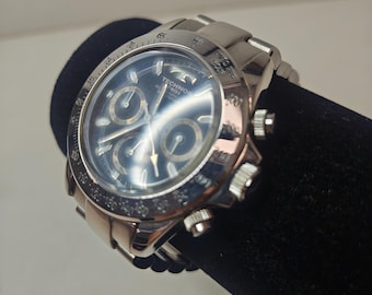 May include: A silver-toned Technos chronograph wristwatch with a dark blue dial, three sub-dials, and a metallic link bracelet. The bezel features a tachymeter scale, and the watch face displays the brand name Technos along with Swiss text.
