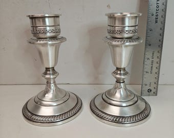 May include: Two silver-toned metal candlesticks featuring a Greek key pattern band below the candle cup and a rope-style border on the circular base. A Westcott stainless steel ruler is positioned vertically, showing a height of approximately 6 inches.
