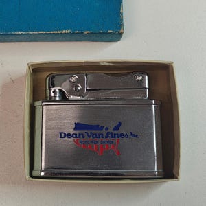 May include: A silver-colored metal lighter with the text "Dean Van Lines, Inc. World Wide Service" printed on the front. The lighter is in a cardboard box with a blue lid.