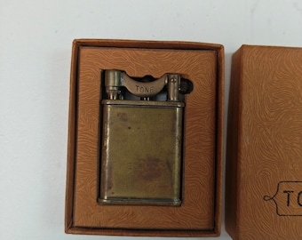 May include: A rectangular brass-toned metal lighter with the word TONE engraved on the top mechanism. The lighter is housed in a brown cardboard box with a textured, swirling pattern and a matching lid featuring the partial text TONE.