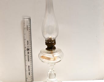 May include: A clear glass oil lamp with a brass burner and a tall, slender chimney. The lamp has a round base and a decorative stem. A ruler is placed next to the lamp. The lamp is approximately 10 inches tall.