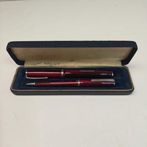 May include: A dark red fountain pen and a matching ballpoint pen set with silver-toned clips and accents, presented in a rectangular navy blue hinged case with a dark blue velvet interior lining and handwritten text on the inner lid.