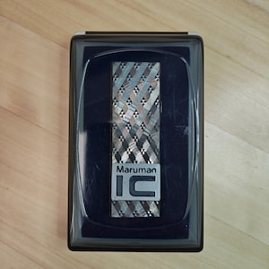 May include: A rectangular lighter with a geometric pattern in shades of silver, grey, and brown, encased in a dark blue plastic box. The lighter has the text "Maruman IC" in silver lettering.