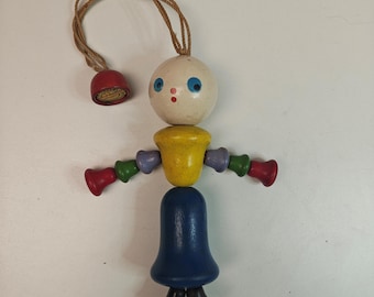 May include: A wooden toy figure featuring a round cream-colored head with blue eyes and a red mouth, a yellow torso, and a blue skirt. The arms and feet are made of small, colorful bell-shaped wooden pieces in red, green, and purple, with a hanging cord.