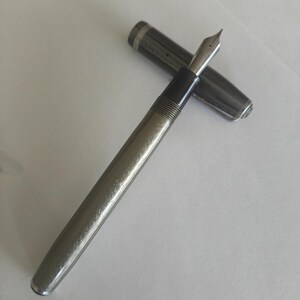 May include: A metallic silver fountain pen with a textured barrel and a dark grip section. The pen features a polished silver-colored nib and a matching detachable cap resting beside it, both displaying a subtle, intricate engraved pattern.