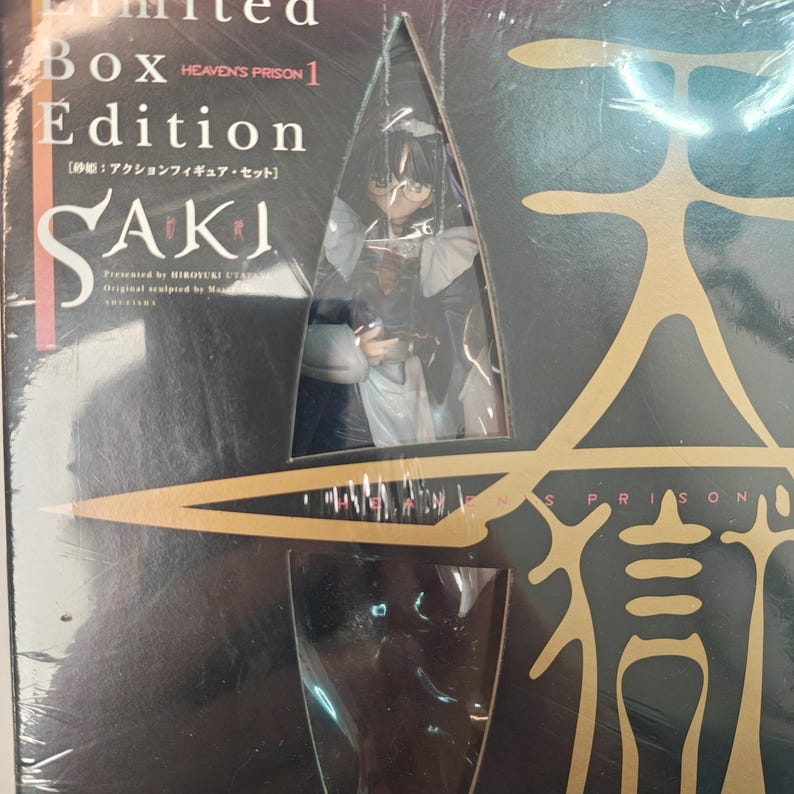 Puede incluir: Un set de caja de edici&oacute;n limitada con una figura de acci&oacute;n SAKI. La caja es negra con detalles dorados y el texto "HEAVEN'S PRISON 1 Edition". La figura es visible a trav&eacute;s de una ventana transparente, mostrando un personaje con ropa oscura.