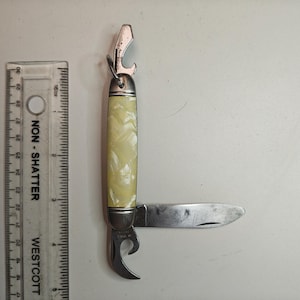 以下が含まれることがあります： クリーム色のハンドルとステンレス鋼の刃と栓抜きが付いた、ヴィンテージのマルチツール。このツールには、缶切りと小さなナイフの刃が含まれています。ハンドルは真珠光沢のある大理石模様です。定規がツールの隣にあります。